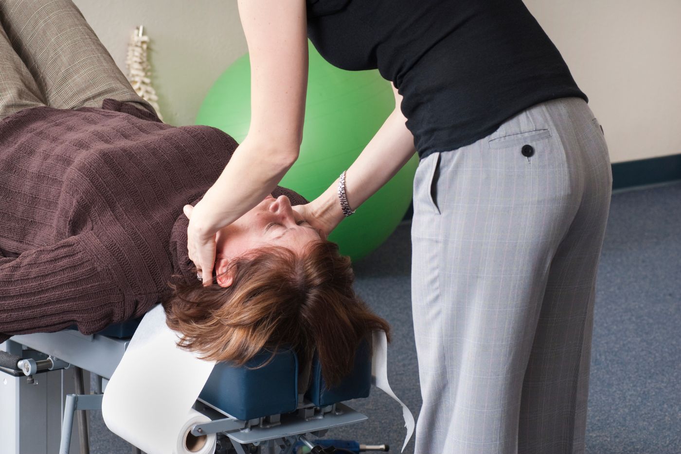 A chiropractor in gray pants adjusts the neck of a female patient lying face down on a specialized therapy bed, with a green exercise ball in the background. A chiropractor in gray pants adjusts the neck of a female patient lying face down on a specialized therapy bed, with a green exercise ball in the background.