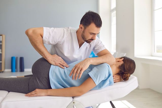 A chiropractor in a white shirt is adjusting a female patient lying on her side on a treatment table in Manhattan Chiropractic Center with a bright window.