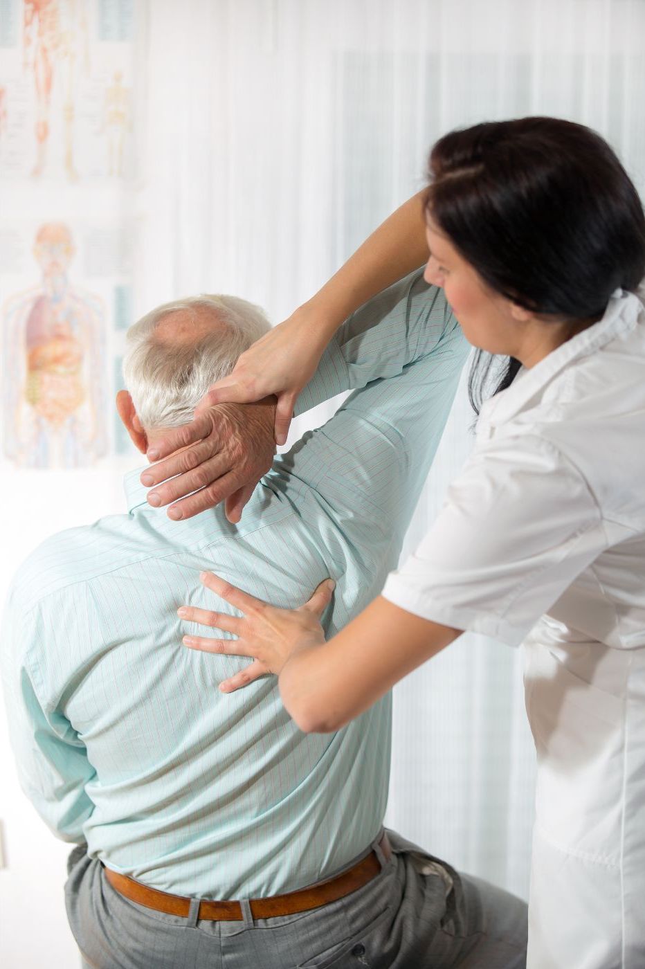 A female chiropractor in a white coat assists a senior male patient with a neck exercise, placing her hand gently on his back and neck in a clinical setting in Manhattan, NY. A female chiropractor in a white coat assists a senior male patient with a neck exercise, placing her hand gently on his back and neck in a clinical setting in Manhattan, NY.