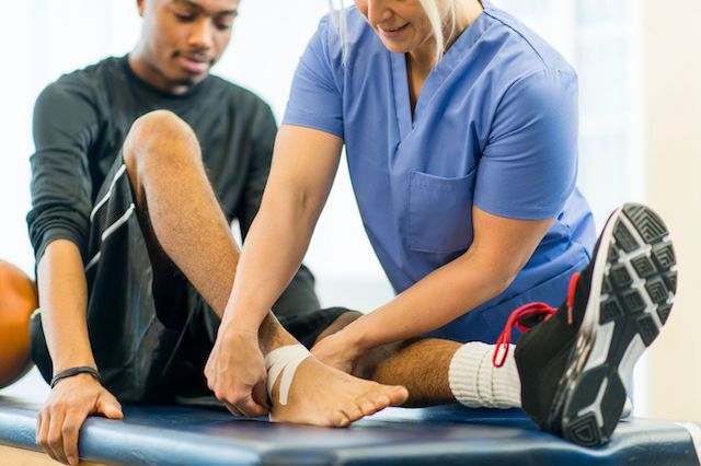 A chiropractor in blue scrubs assists a male athlete with a bandaged ankle during a rehabilitation session in a clinic setting.
