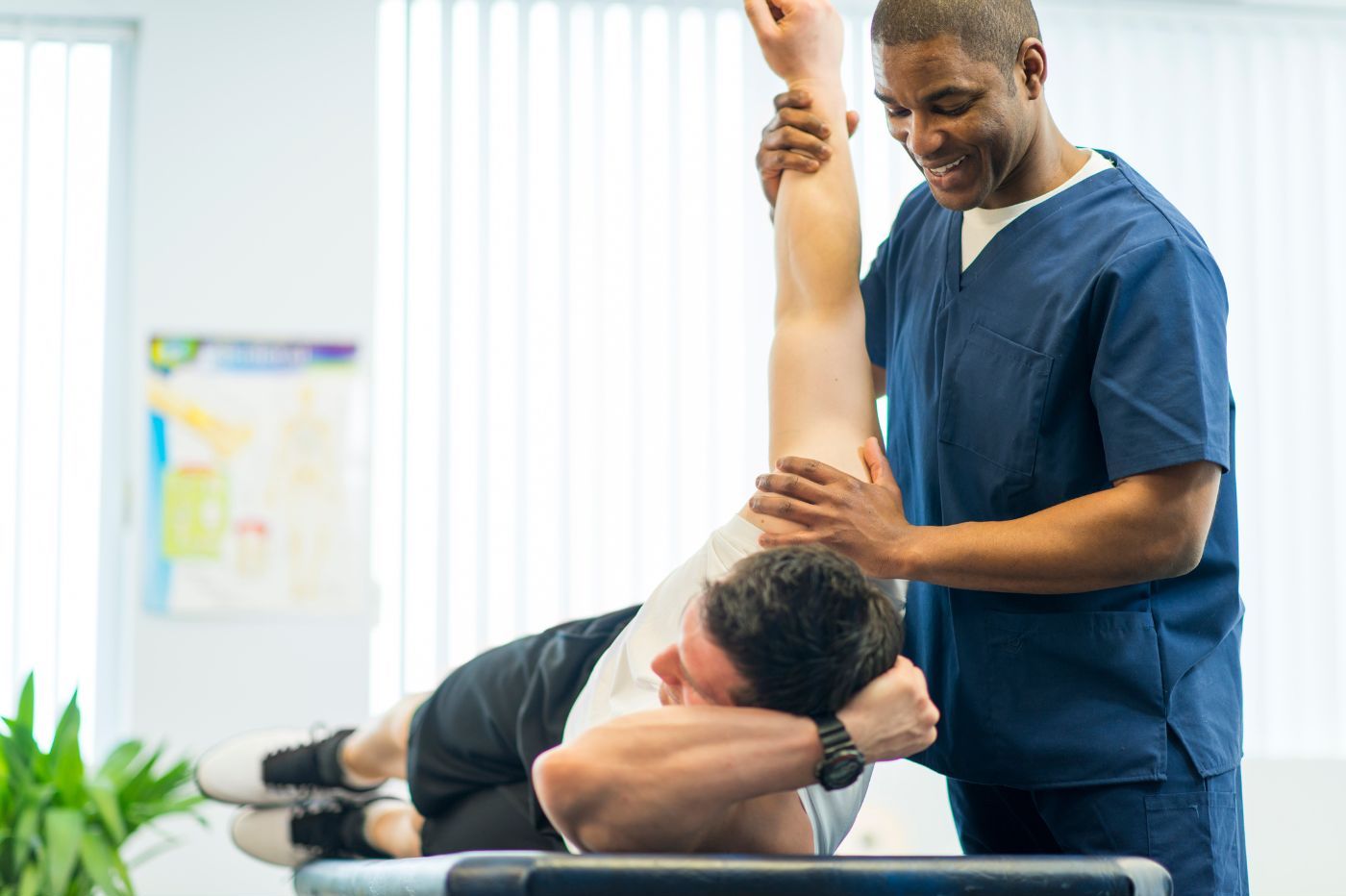 A chiropractor, a black man in blue scrubs, assists a white male patient performing a stretching exercise on a medical table in a brightly-lit clinic. A chiropractor, a black man in blue scrubs, assists a white male patient performing a stretching exercise on a medical table in a brightly-lit clinic.