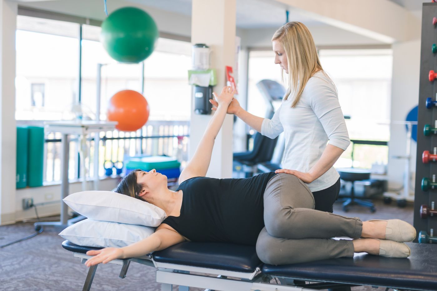 A chiropractor assists a female patient performing a leg stretch on a therapy table in Manhattan Chiropractic Center with exercise balls in the background. A chiropractor assists a female patient performing a leg stretch on a therapy table in Manhattan Chiropractic Center with exercise balls in the background.