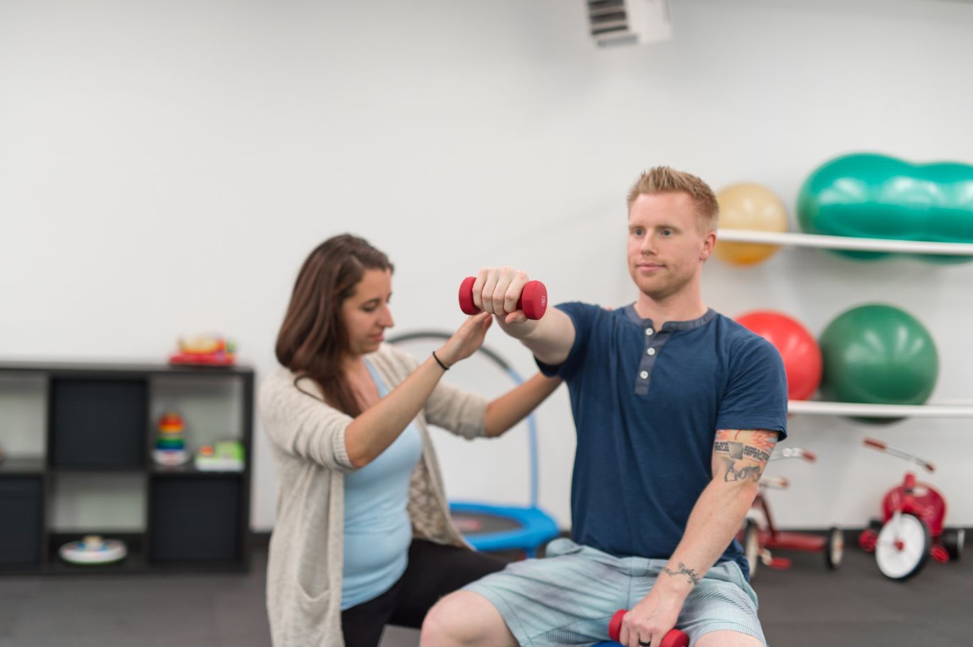 A chiropractor assisting a young man with arm rehabilitation using a red dumbbell in a Manhattan Chiropractic Center clinic equipped with various exercise equipment and colorful balls in the background. A chiropractor assisting a young man with arm rehabilitation using a red dumbbell in a Manhattan Chiropractic Center clinic equipped with various exercise equipment and colorful balls in the background.