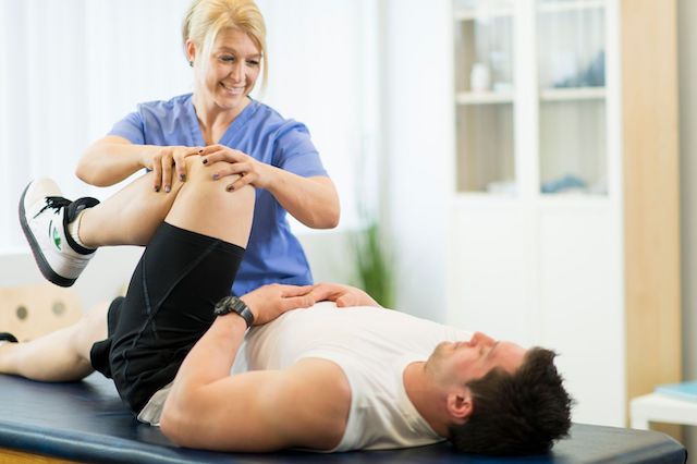 A physical therapist assists a male patient with a leg exercise, both smiling, in a bright clinic setting.