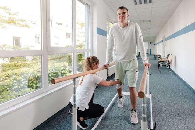 A young man using crutches smiles while walking in a hallway at Manhattan Chiropractic Center with assistance from a female therapist, who holds a wooden bar for support.