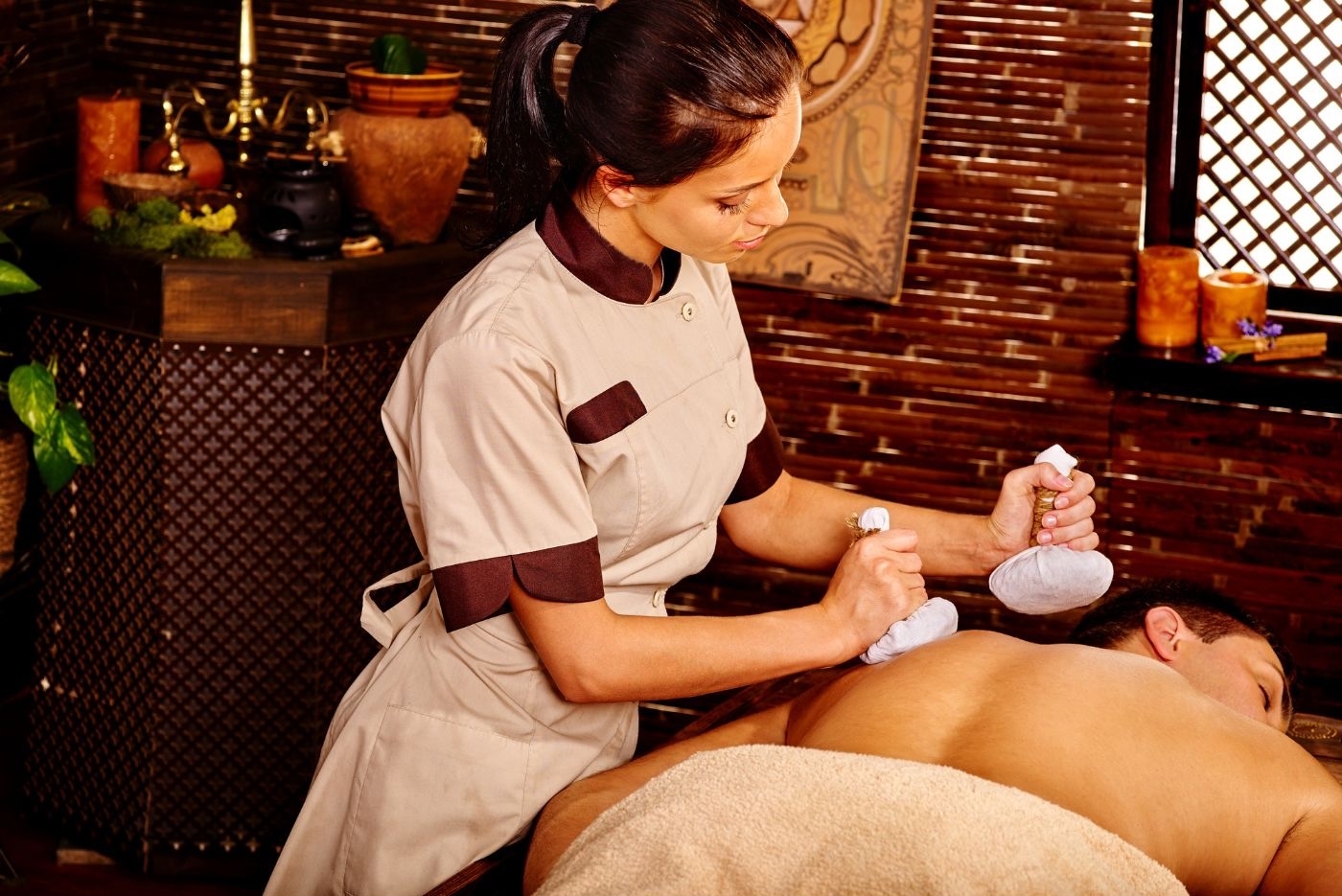 A massage therapist in Manhattan uses herbal compress balls on the back of a client in a spa setting. The therapist wears a beige and brown uniform, and the room is decorated with wooden elements, plants, and warm lighting, creating an ambiance reminiscent of Ayurvedic medicine techniques. A massage therapist in Manhattan uses herbal compress balls on the back of a client in a spa setting. The therapist wears a beige and brown uniform, and the room is decorated with wooden elements, plants, and warm lighting, creating an ambiance reminiscent of Ayurvedic medicine techniques.