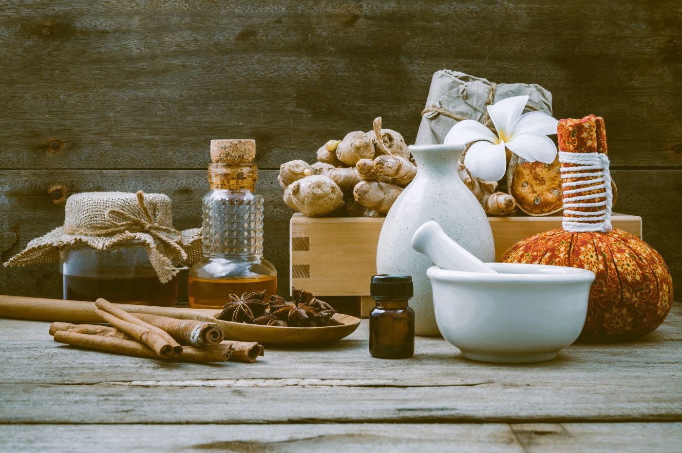 A rustic arrangement of spa and wellness items on a wooden table. Includes a mortar and pestle, essential oil bottles, jars wrapped in burlap, cinnamon sticks, star anise, dried herbs reminiscent of Ayurvedic Medicine, and a tropical flower in a ceramic vase. A wooden backdrop completes the scene. A rustic arrangement of spa and wellness items on a wooden table. Includes a mortar and pestle, essential oil bottles, jars wrapped in burlap, cinnamon sticks, star anise, dried herbs reminiscent of Ayurvedic Medicine, and a tropical flower in a ceramic vase. A wooden backdrop completes the scene.