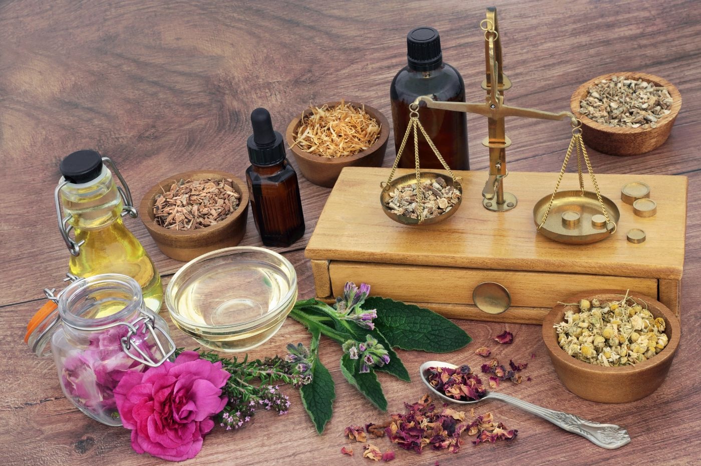 A wooden table in a Manhattan studio displays various traditional herbal medicine items, including bottles of essential oils, dried herbs in bowls, fresh flowers, a spoonful of dried petals, and a vintage balance scale. The scene evokes the naturopathic approach to wellness favored by NYC's holistic community.
