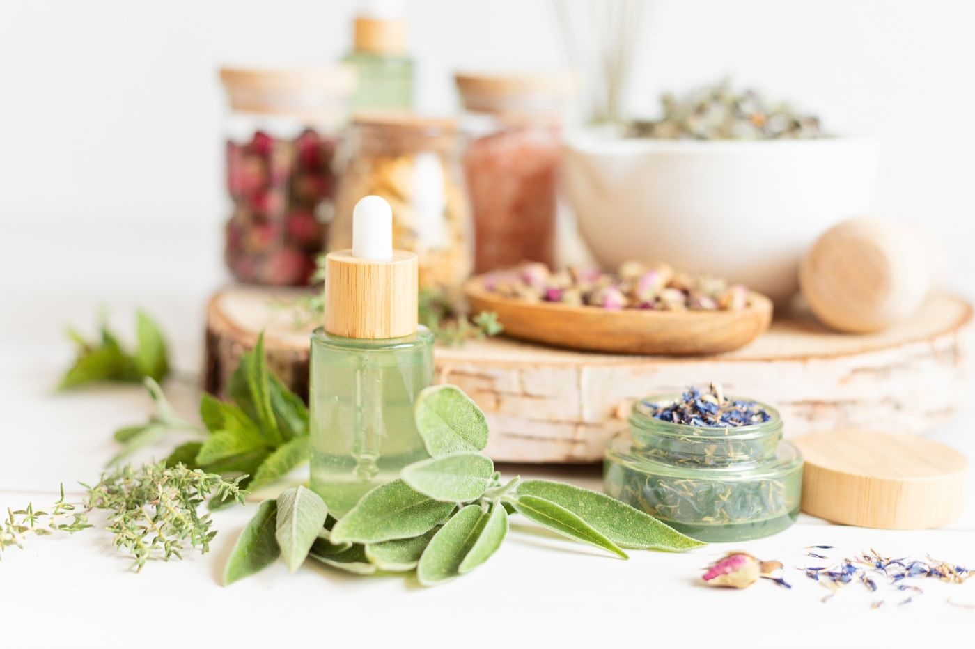 A collection of natural skincare items on a white surface, including a dropper bottle with green liquid, dried herbs, flowers, and leaves in glass jars, and small wooden bowls filled with dried botanical ingredients. A round wooden cutting board is in the background—perfect for naturopathic doctors in NYC.