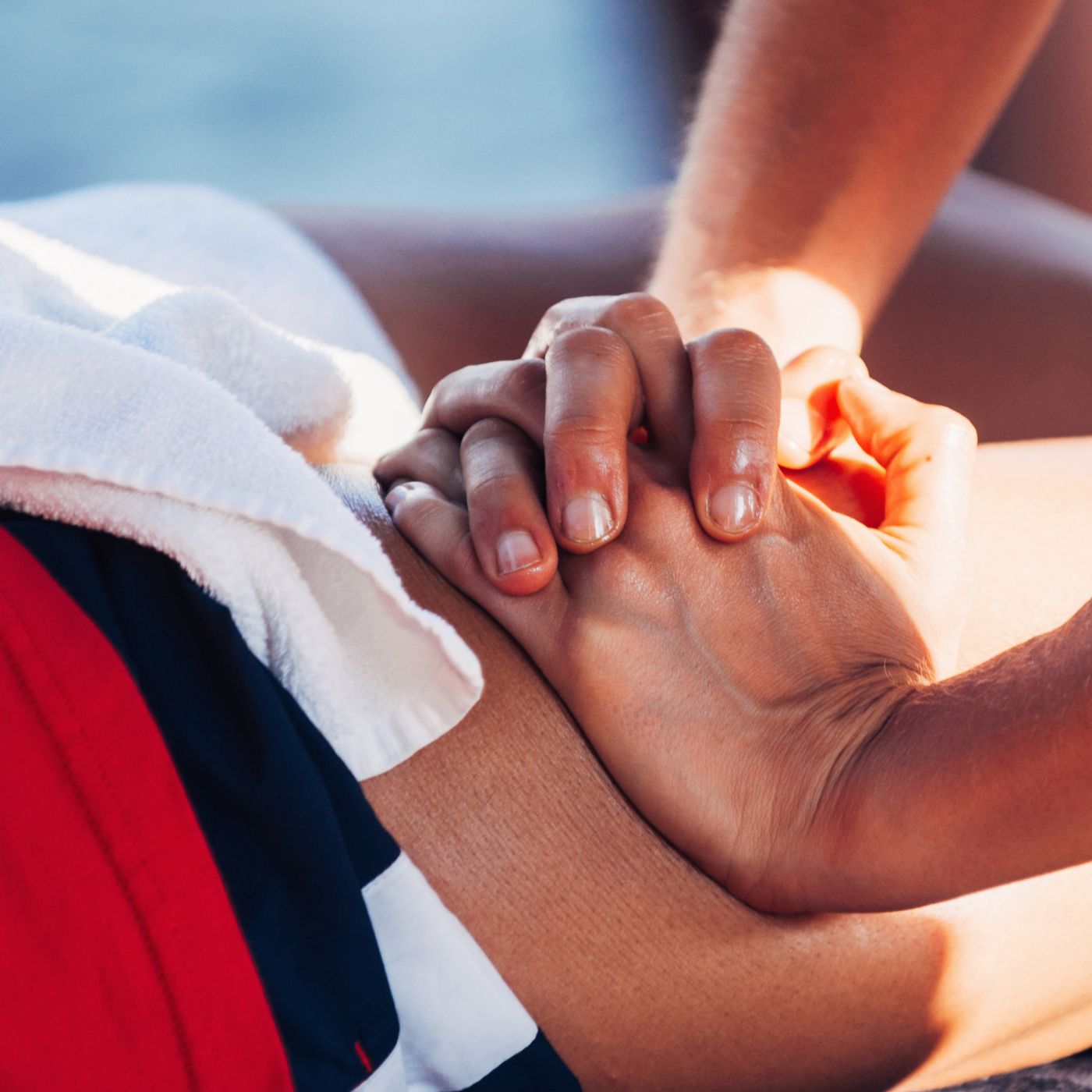 Close-up of a person performing chest compressions on another individual during CPR in Manhattan. The hands are interlocked and pressing down on the chest of a person lying on a surface with a white towel on their side. Sunlight shines on the scene. Close-up of a person performing chest compressions on another individual during CPR in Manhattan. The hands are interlocked and pressing down on the chest of a person lying on a surface with a white towel on their side. Sunlight shines on the scene.