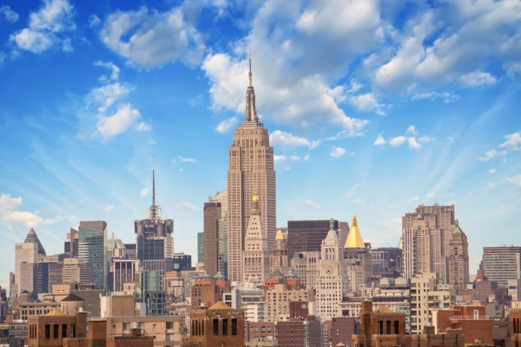 A panoramic view of the midtown Manhattan skyline featuring the Empire State Building under a blue sky with scattered clouds.