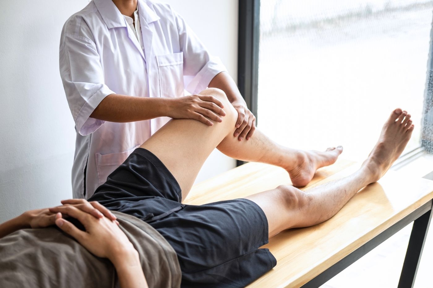A person wearing a white coat is performing a sports physical therapy session on another person's knee. The individual receiving the therapy, dressed in black shorts, is lying on a treatment table. The setting appears to be a bright clinic in Manhattan, NYC. A person wearing a white coat is performing a sports physical therapy session on another person's knee. The individual receiving the therapy, dressed in black shorts, is lying on a treatment table. The setting appears to be a bright clinic in Manhattan, NYC.
