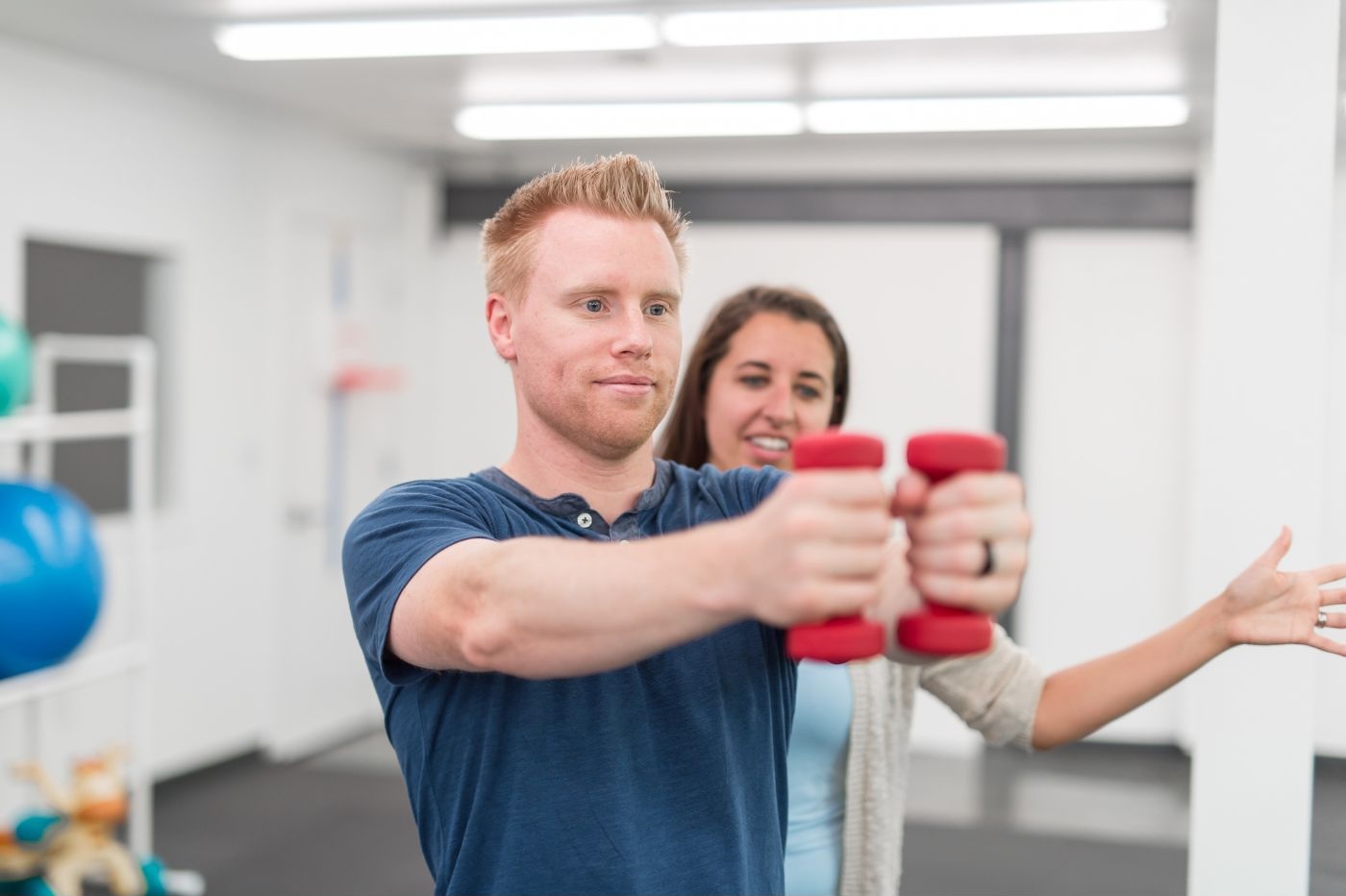 A man in a blue shirt focuses on lifting two red dumbbells, extending his arms forward. A woman in casual attire stands behind him, appearing to instruct or assist with his exercise at a sports physical therapy center in Manhattan. The brightly lit fitness facility reflects the energy of NYC. A man in a blue shirt focuses on lifting two red dumbbells, extending his arms forward. A woman in casual attire stands behind him, appearing to instruct or assist with his exercise at a sports physical therapy center in Manhattan. The brightly lit fitness facility reflects the energy of NYC.