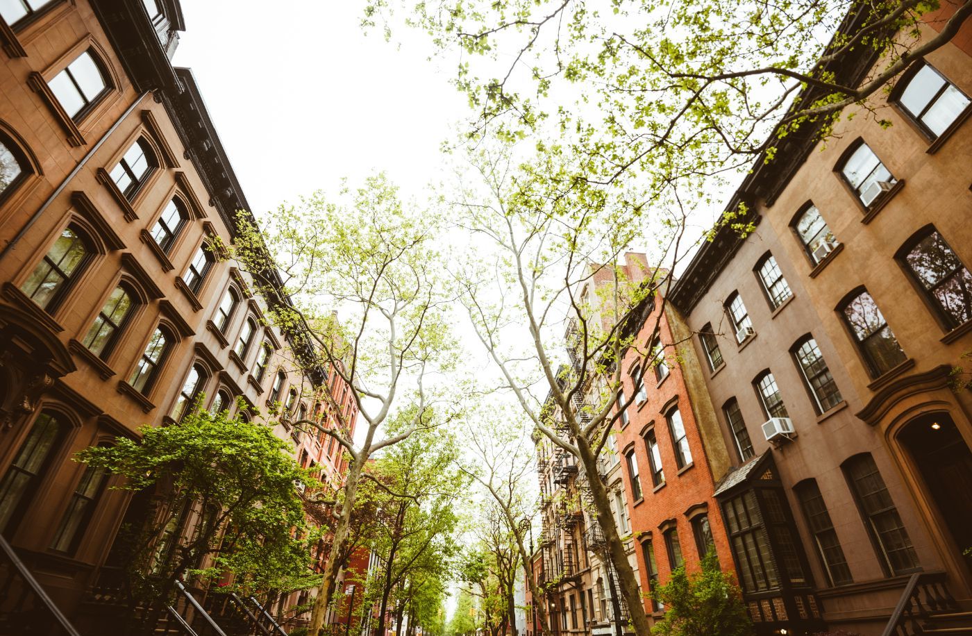 Low-angle view of a tree-lined street with historic brownstone buildings in Upper Manhattan, capturing the essence of a spring day with green leaves and a warm ambiance. Low-angle view of a tree-lined street with historic brownstone buildings in Upper Manhattan, capturing the essence of a spring day with green leaves and a warm ambiance.
